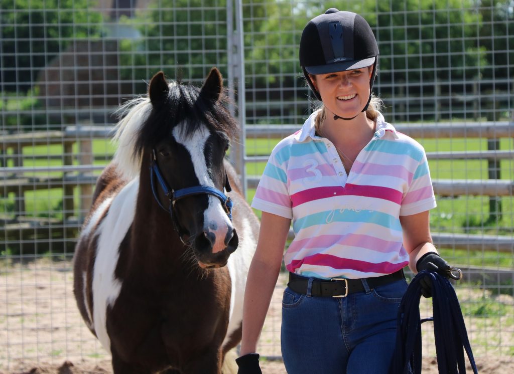 woman wearing a stripy top and a riding helmet, stood next to a skewbald cob