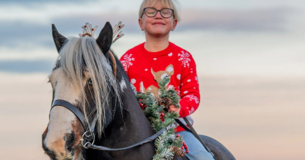 young girl wearing a christmas jumper sat atop a cob with a wreath around his neck