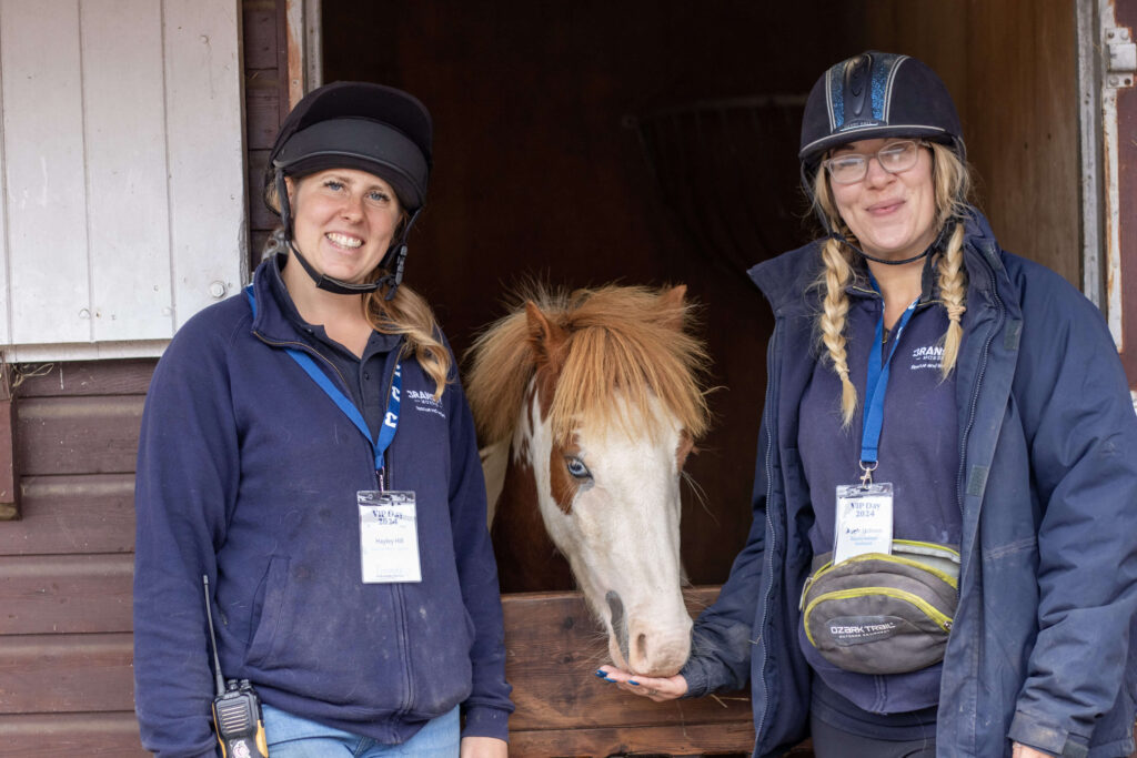 two members of staff stood either side of a stable, a skewbald pony is poking his head over the door