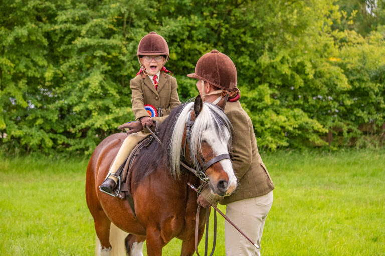 Peyton smiling proudly while riding Skip, holding a rosette, with an adult beside her in matching equestrian attire. They are in a grassy field with trees in the background