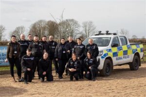 Group of Nottinghamshire Police rural crime officers and Bransby Horses staff standing and kneeling together in front of a marked Rural Police vehicle, outdoors on a sandy area with trees and a hedgerow in the background.