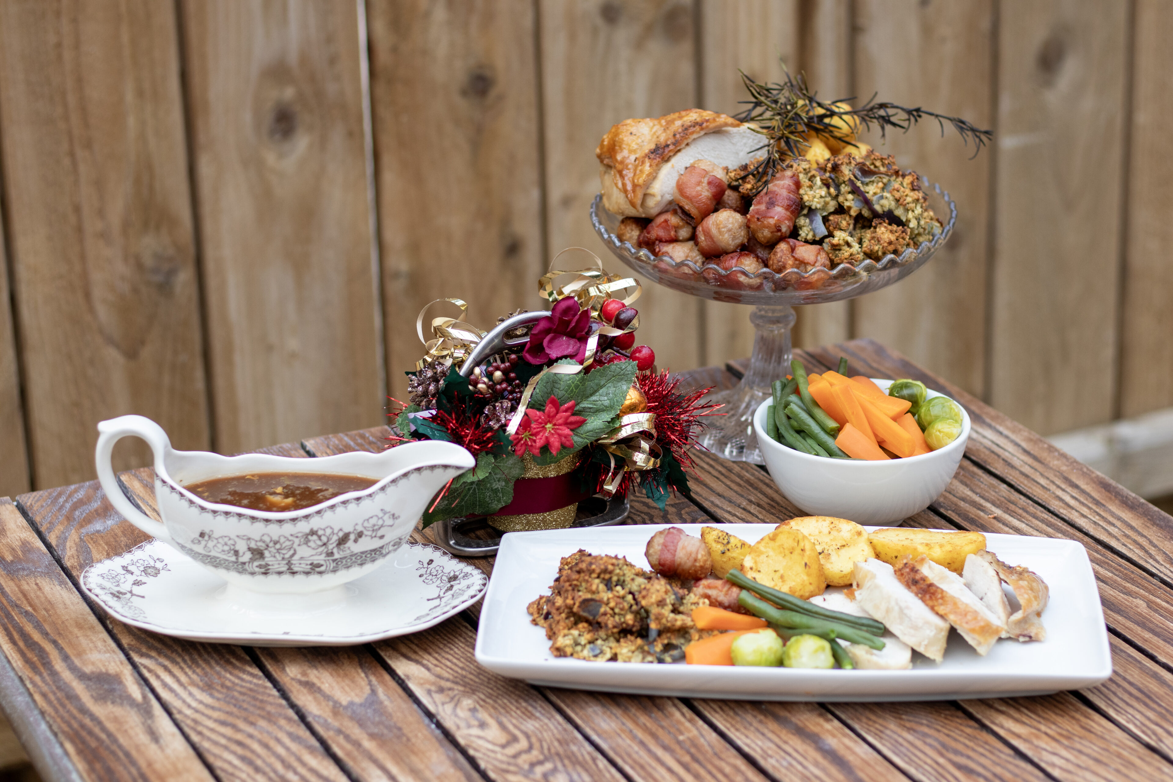 A festive Christmas dinner at Bransby Horses featuring a plate of roast turkey slices, golden roast potatoes, Brussels sprouts, green beans, carrots, and stuffing. In the background, a raised platter holds extra stuffing, pigs in blankets, and roasted meat garnished with rosemary. A decorative gravy boat sits to the side, and a small Christmas centerpiece with red flowers and gold accents adds a seasonal touch